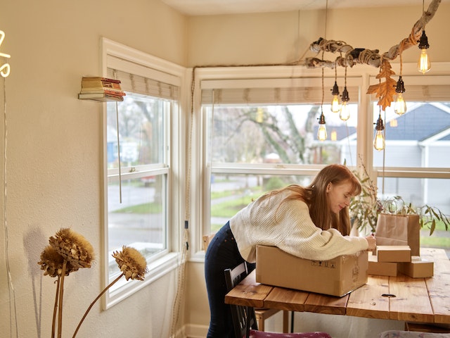 A woman packing a box on a wooden table by a window.